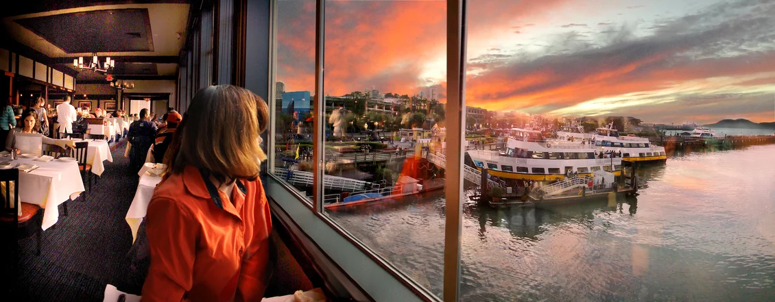 Woman dining by a window at Fog Harbor, overlooking boats and a vibrant sunset at San Francisco’s PIER 39.